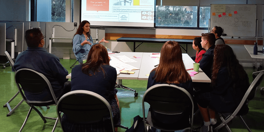 A group of students sit around a table with a researcher speaking to them.,