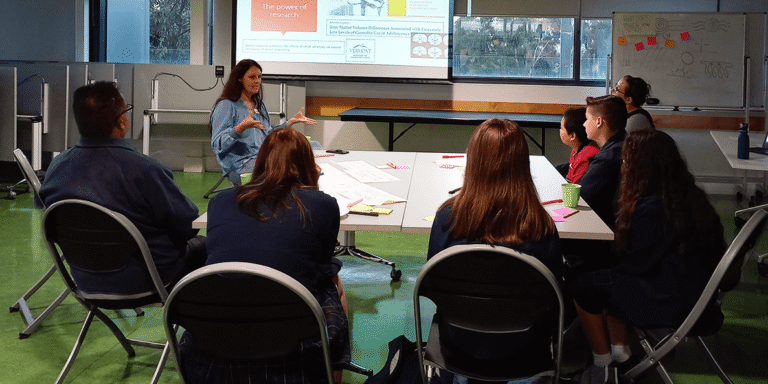 A group of students sit around a table with a researcher speaking to them.,