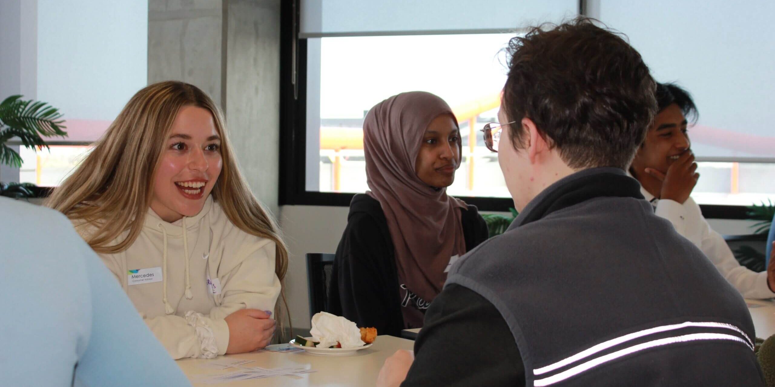 A girl with blonde hair smiles at a boy sitting across the table from her. Next to her is another girl with a purple hijab.