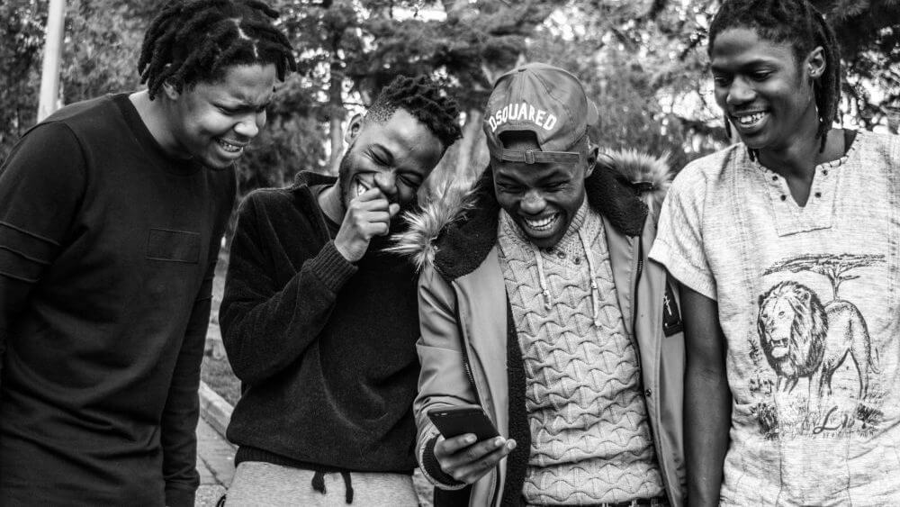 Black and white photo of four young african men looking at a phone and laughing