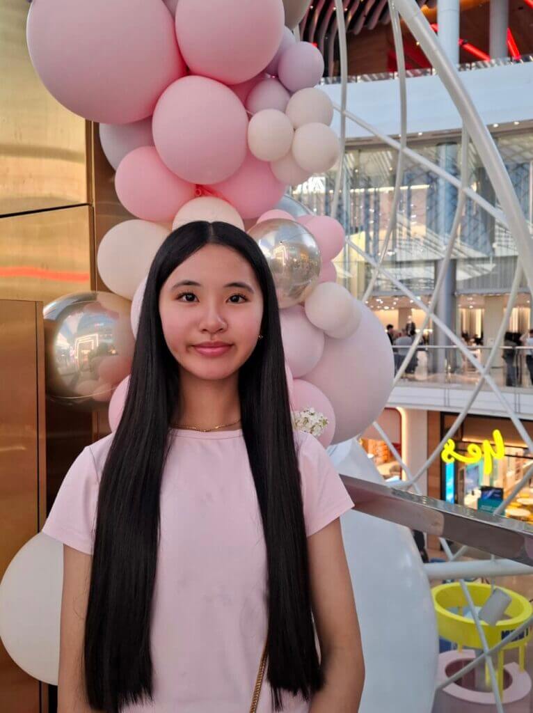 Jayda is wearing a pink shirt and smiling at the camera. Behind her there is a pink and silver balloon sculpture.