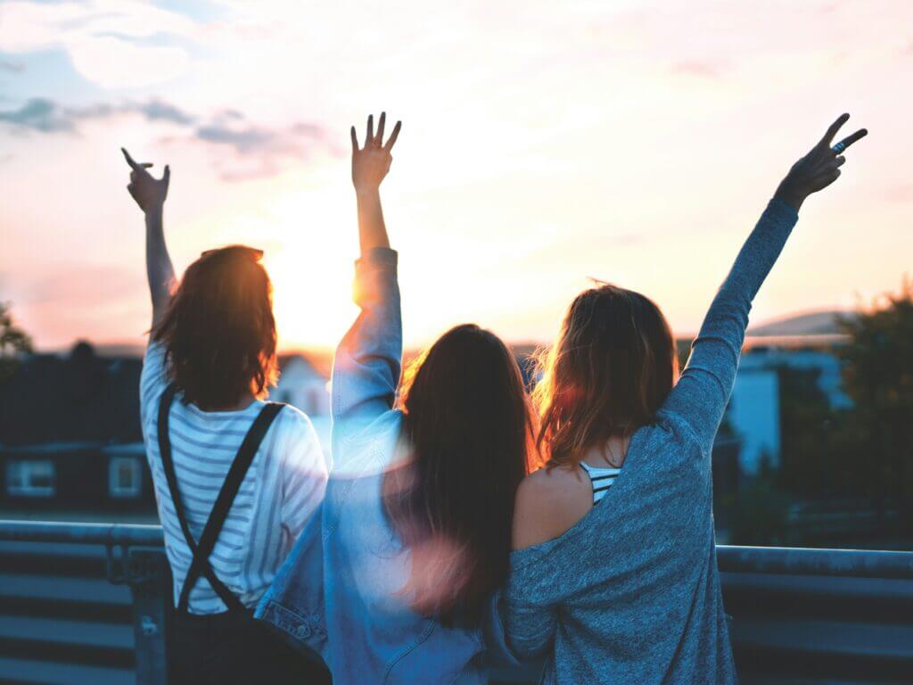 Three women face their backs to the camera, looking at a sunset with their arms in the air