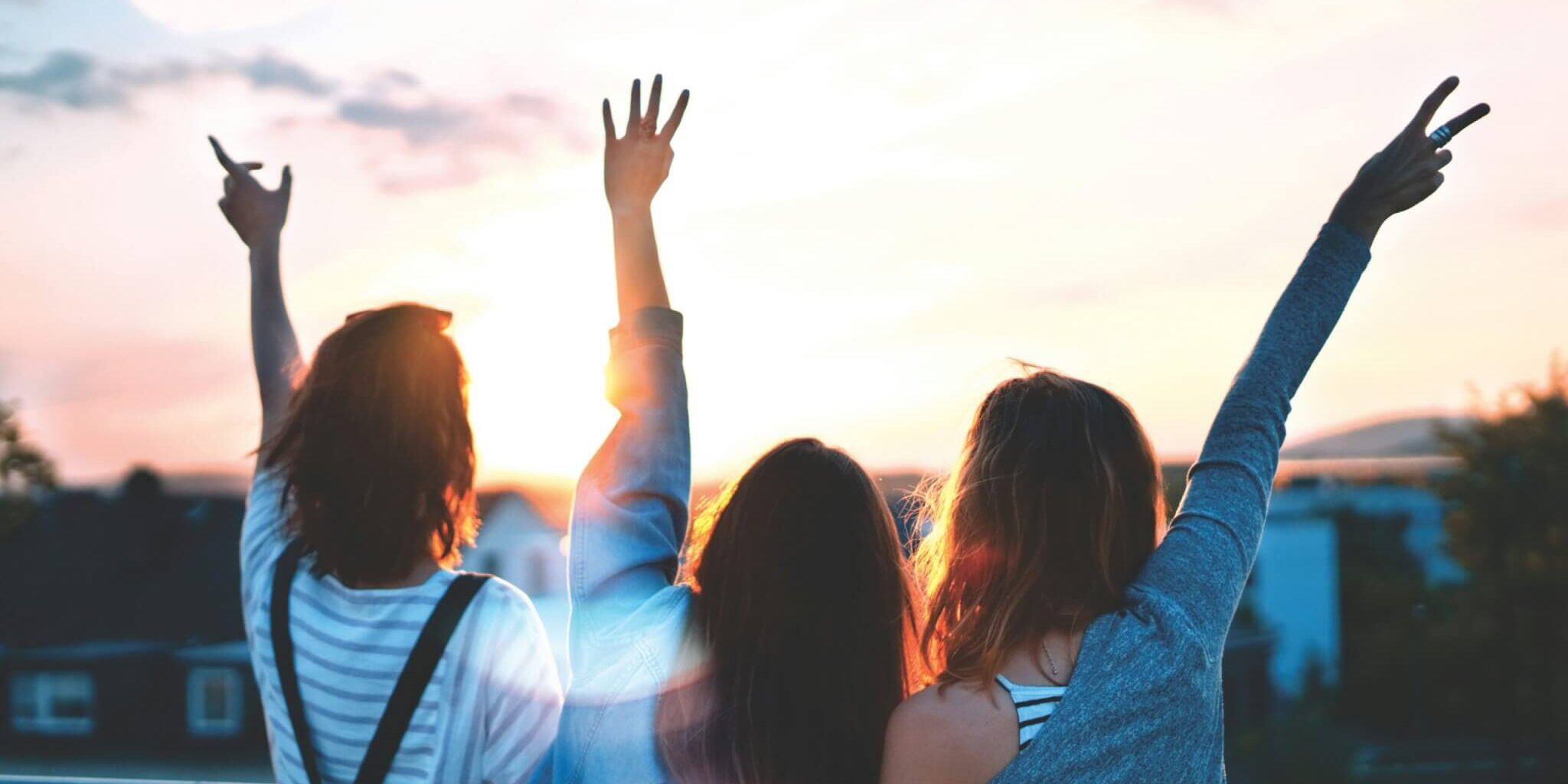 Three women face their backs to the camera, looking at a sunset with their arms in the air