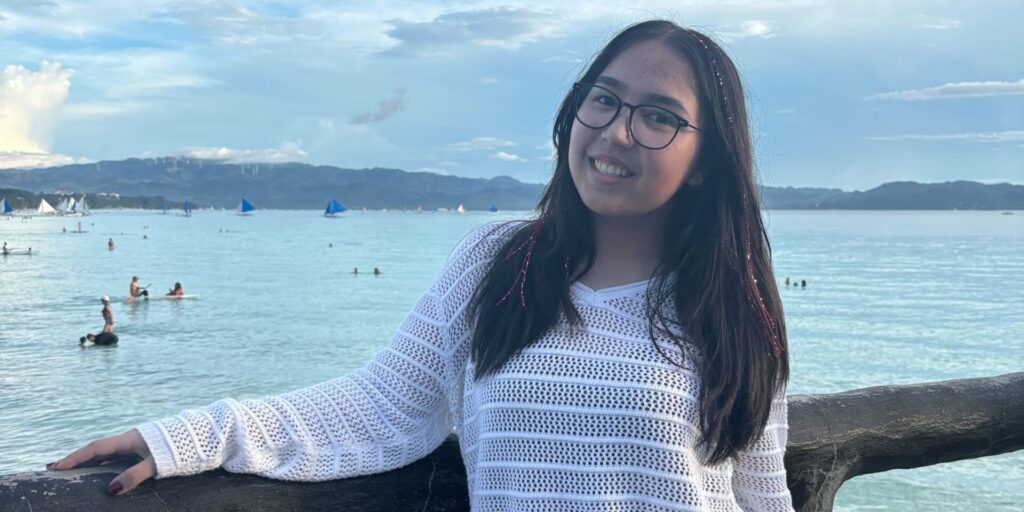 Zoe is wearing a striped long sleeve and leaning against railing. The ocean is behind her.