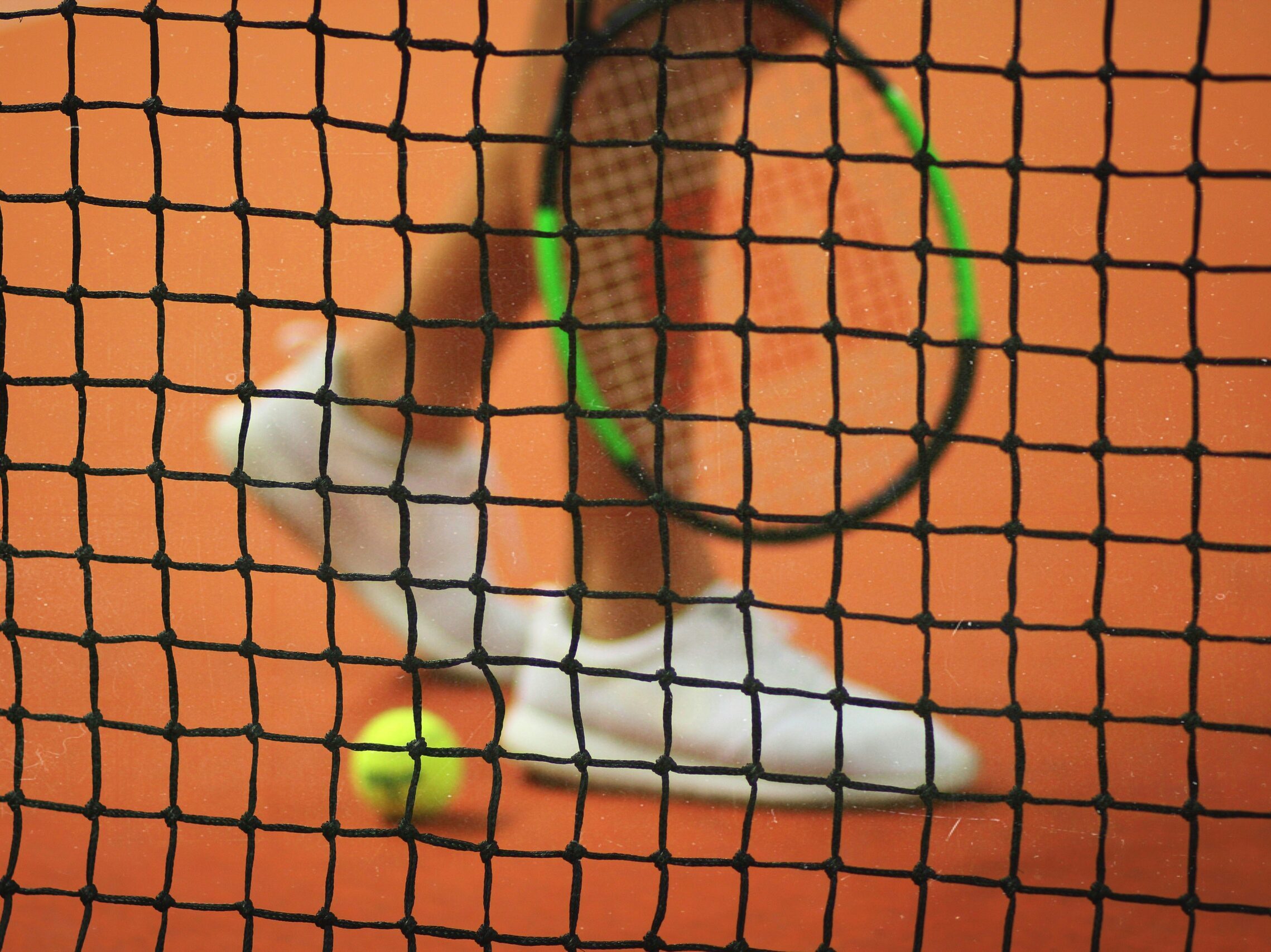 Shot through a tennis court net, there is a blurry closeup of legs, with a tennis ball on the ground and a tennis racquet