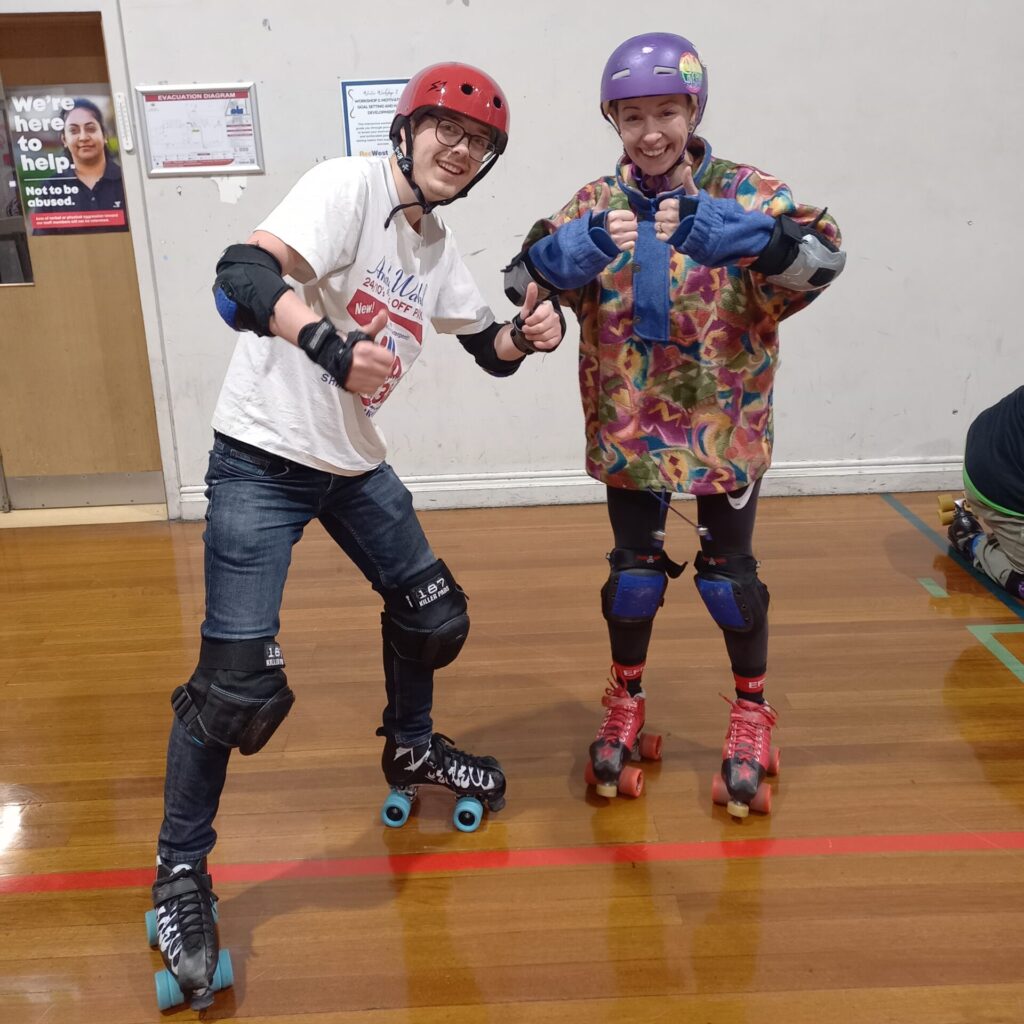 Two people wearing helmets and roller skates smile at the camera with their thumbs up