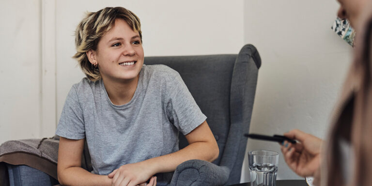 A young person sits in a chair smiling and talking to someone not visible in the frame