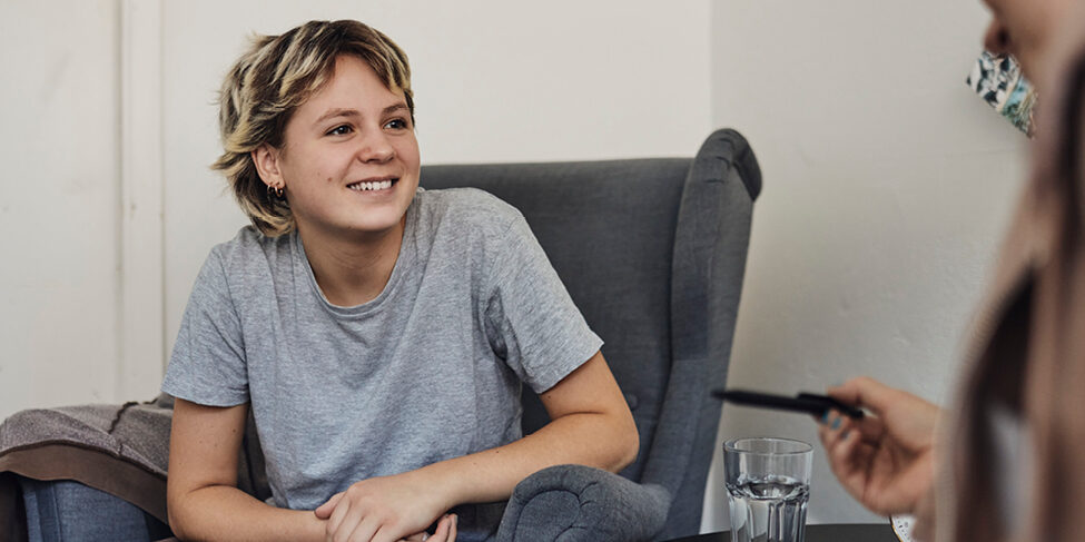 A young person sits in a chair smiling and talking to someone not visible in the frame