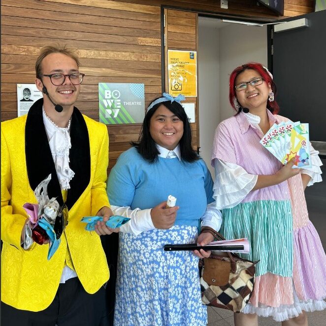 Three people wear bright colourful clothing and smile at the camera while holding brochures