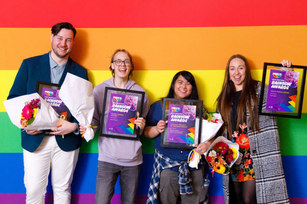 A group of four people hold framed certificates and flowers while smiling at the camera with a rainbow flag background