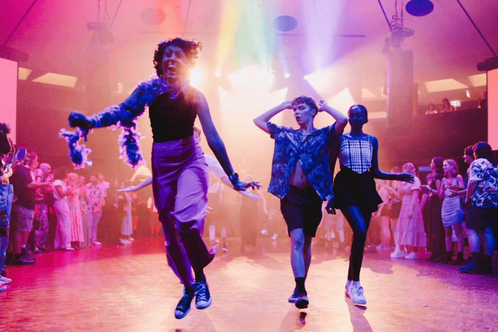 Three people skip across a dance floor, covered in pink and purple light